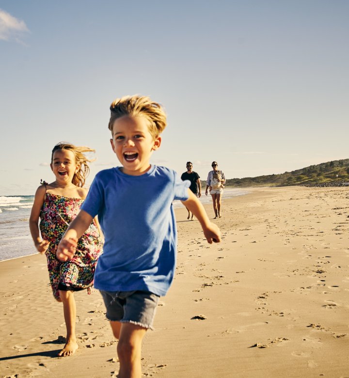 two children running on beach with parental figures in the background