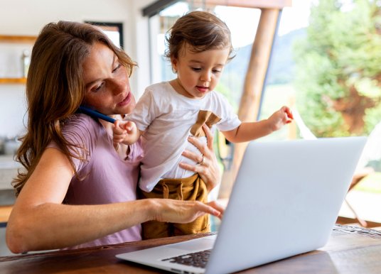 Woman sitting at a table trying to talk on a mobile whilst holding a child and typing on a laptop