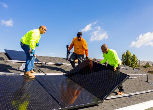 Three workers installing large solar panels on the roof of a house.