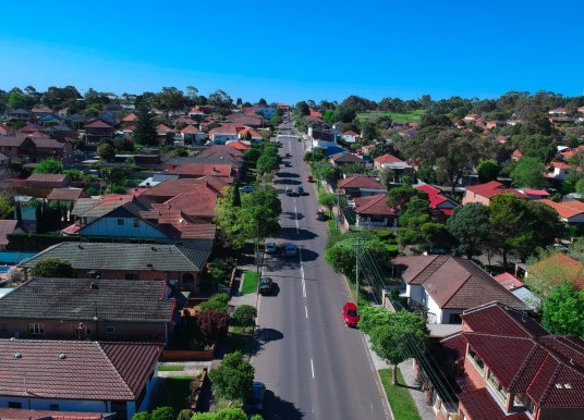 An image of lots of houses in the suburb of a major city or town, with a road running through the middle of the houses. Adding to the sense of an Australian suburban scene is bright blue sky over the horizon.